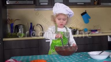 一个严肃的小女孩，用勺子把面团混合在厨房的玻璃碗里
